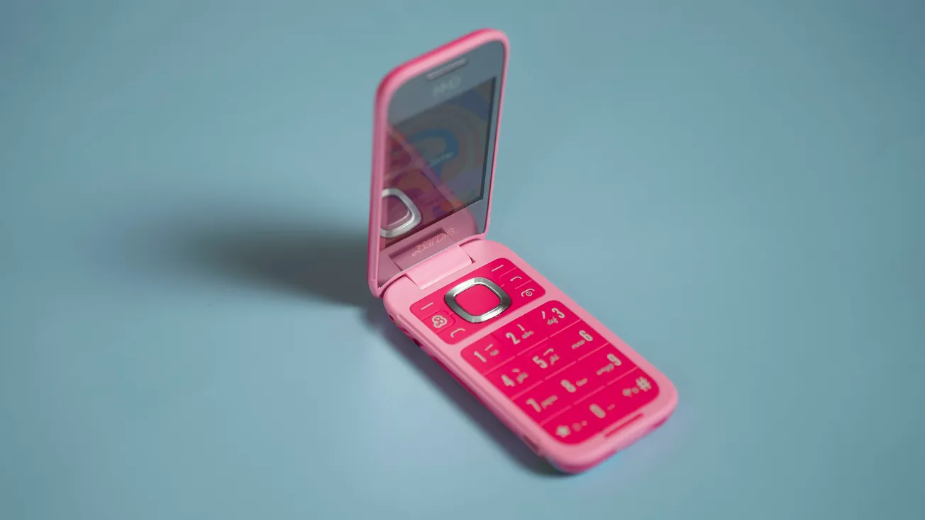 A pink flip phone is open on a light blue surface, displaying large buttons and a small internal screen. The tone is nostalgic and retro.