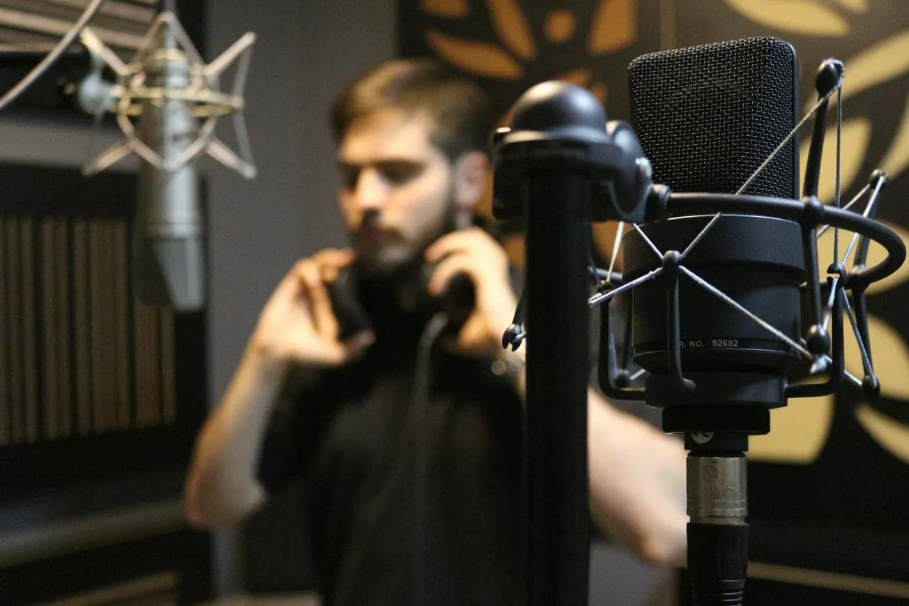 A focused microphone in a recording studio, with a blurred person adjusting headphones in the background, creating a professional, creative atmosphere.