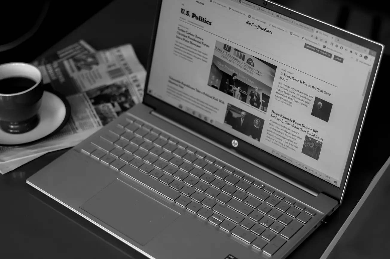Black and white image of a laptop on a table displaying U.S. politics news. Nearby, a cup of coffee and scattered newspapers suggest a serious, focused ambiance.