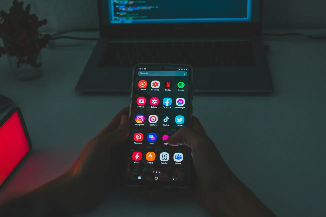 Hands holding a smartphone displaying colorful app icons in a dim room, with a laptop in the background, creating a focused and tech-savvy atmosphere.