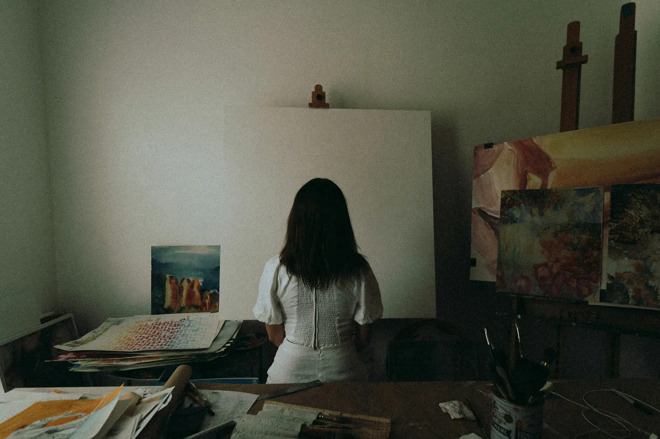 A woman in a white dress stands in front of a blank canvas in a dimly lit art studio. Surrounding her are colorful paintings and art supplies.