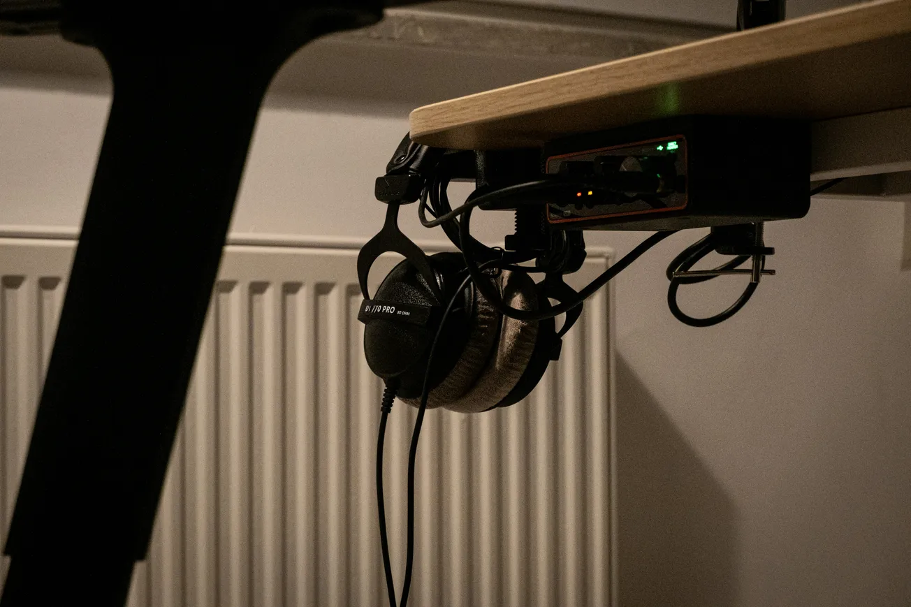 Headphones hanging below a wooden desk, next to a small electronic device with glowing green lights. The setting has a cozy, dimly lit ambiance.