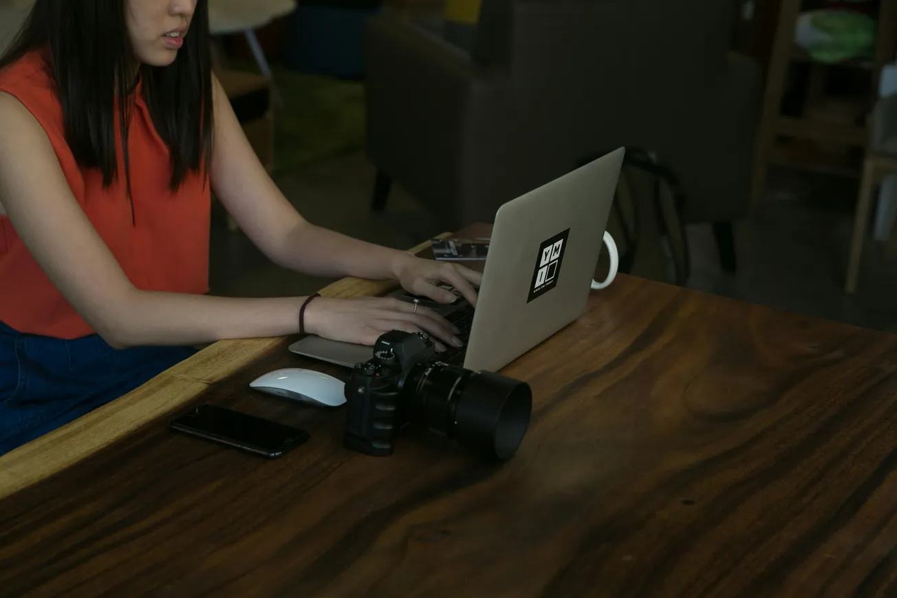 A woman in a red top uses a laptop at a wooden table, alongside a camera, smartphone, and mouse in a cozy, modern setting, conveying focus and creativity.