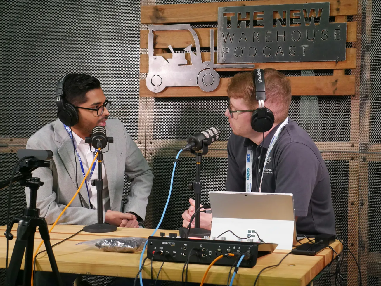 Two men wearing headphones and speaking into microphones are seated at a wooden table for "The New Warehouse Podcast" recording.