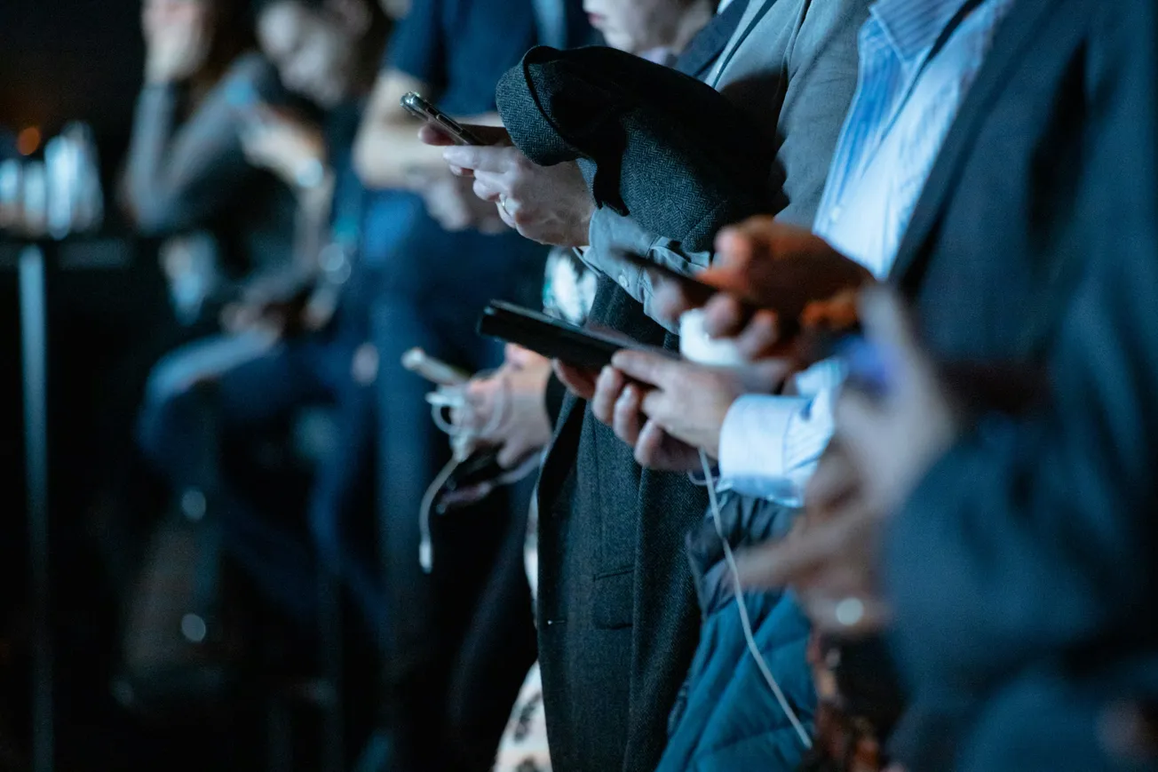 A group of people in business attire stand closely, engrossed with their smartphones. The scene is dimly lit, conveying a focused, absorbed atmosphere.