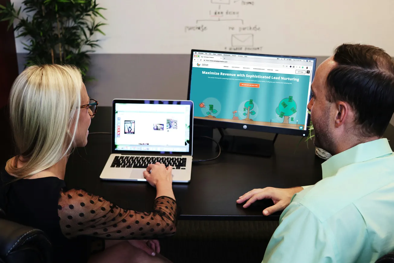 Two colleagues discuss marketing strategies in an office. One points to a laptop screen, while the other looks at a desktop monitor showing a lead nurturing webpage.
