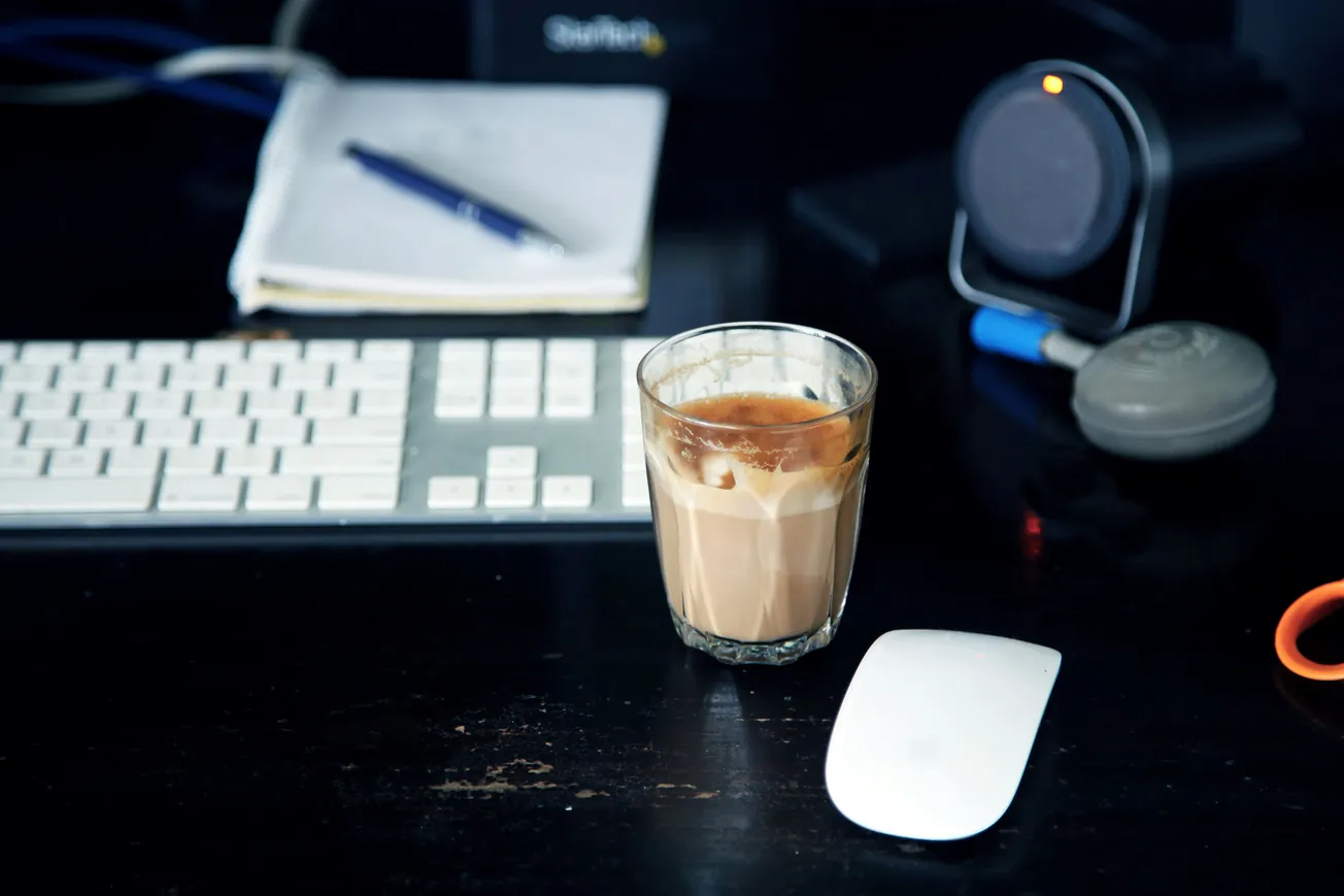 A glass of coffee sits on a dark desk, surrounded by a white keyboard, mouse, notebook, and speaker, creating a cozy, productive ambiance.