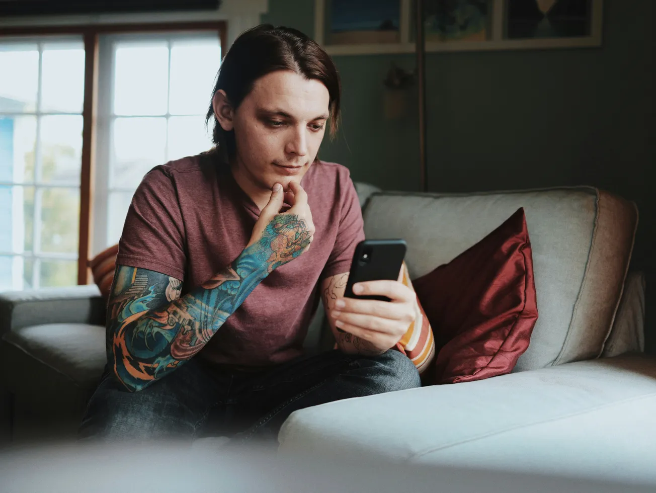 A man with colorful tattoo sleeves sits on a beige sofa, focused on his smartphone. He's wearing a maroon t-shirt, with soft daylight filtering through a window.