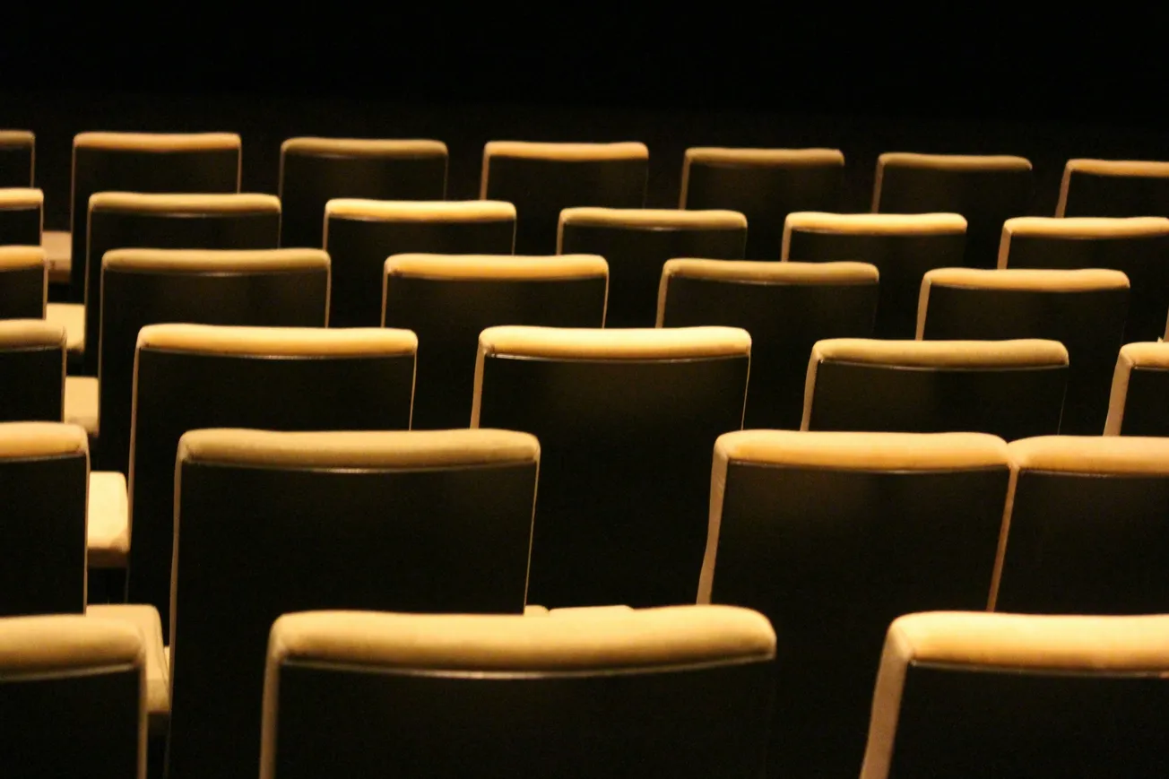 Empty theater seats with dim, warm lighting create a calm, anticipatory atmosphere. Rows of evenly spaced chairs face a dark, unseen stage.