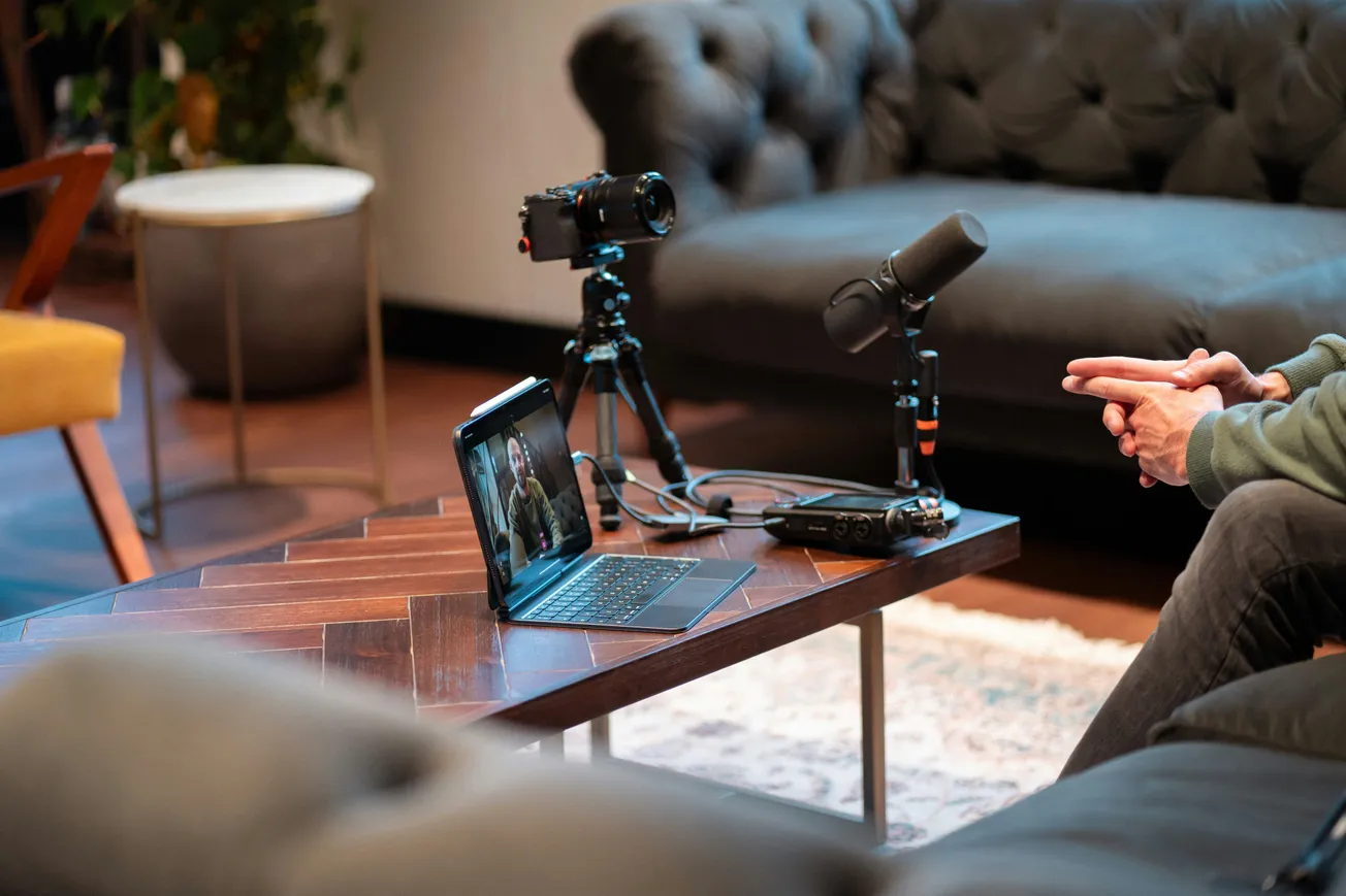 Cozy living room setup for a video podcast, featuring a laptop, camera, and microphone on a wooden table. A person gestures while speaking.