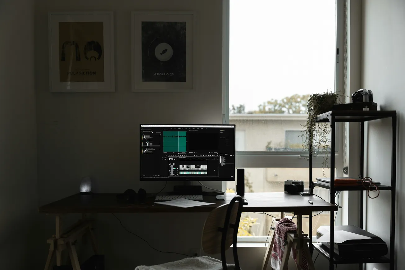 A dimly lit home office with a computer monitor displaying video editing software. A window shows an overcast sky. Minimalist decor with a calm atmosphere.