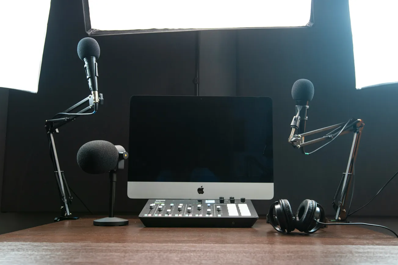 A podcast setup with a computer at the center, two microphones on adjustable arms, a mixing console, and headphones. Bright lighting creates a professional vibe.