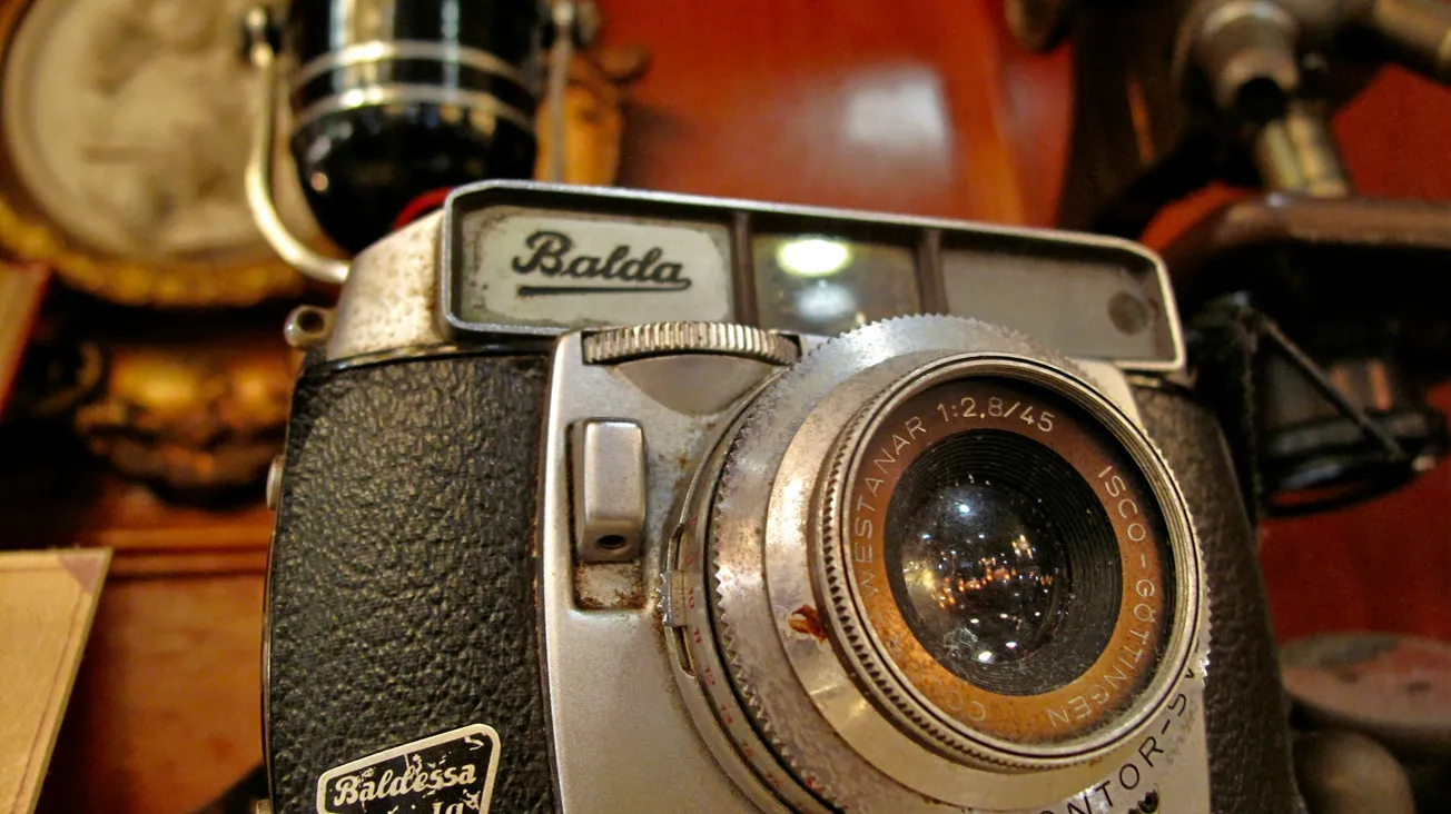 Close-up of a vintage Balda camera with a detailed lens, set against a blurred background of antique items, evoking nostalgia and history.