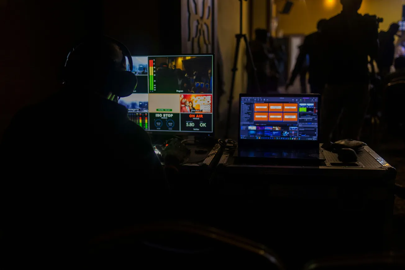 A person wearing headphones sits in a dimly lit room, operating video production equipment. Two monitors display colorful graphics and broadcast controls.