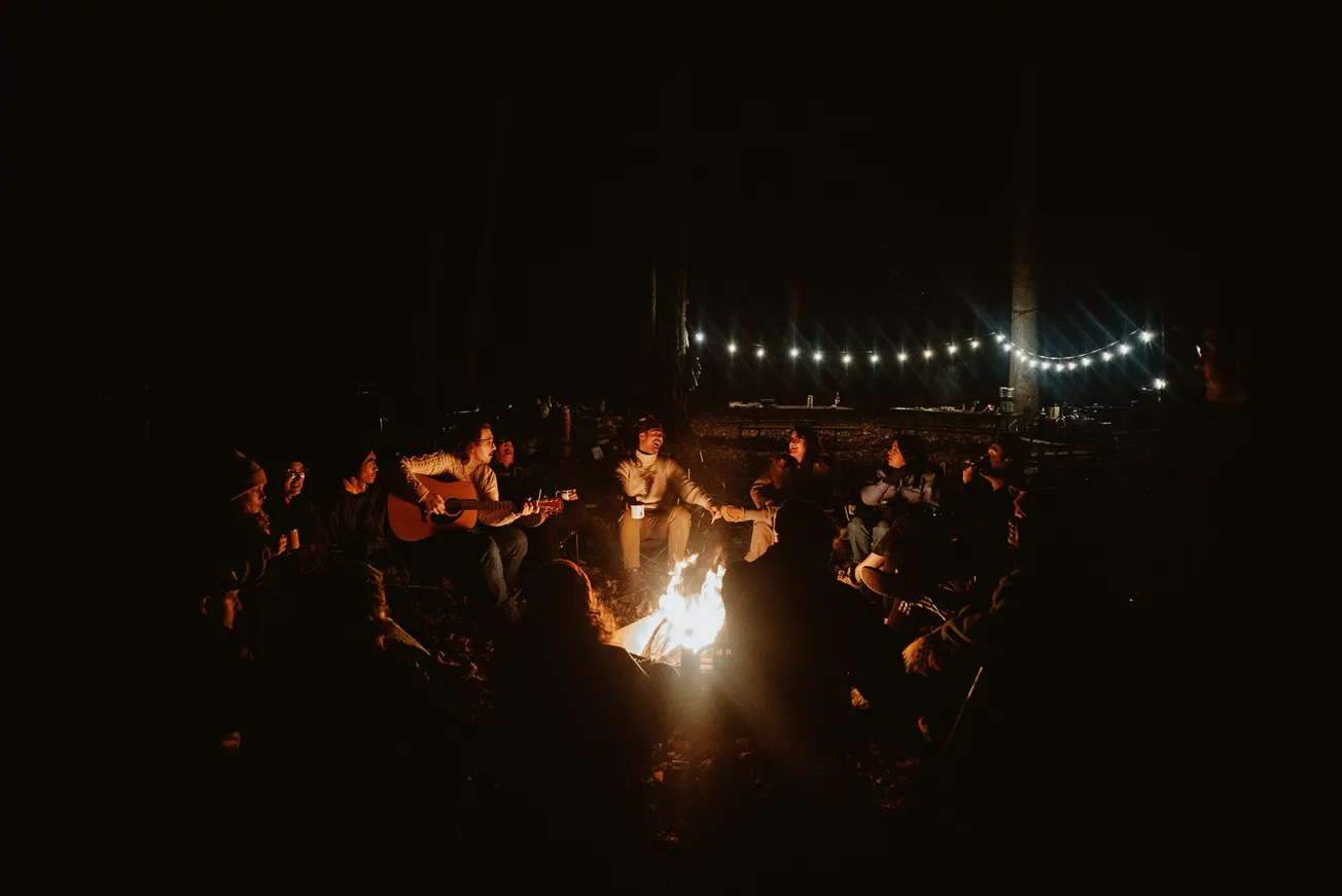 A group of people sit around a campfire at night, with one playing guitar. String lights hang in the background, creating a warm, cozy atmosphere.
