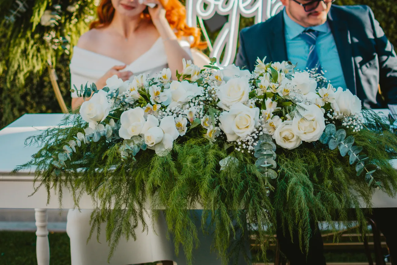 A couple sits behind a table adorned with lush greenery and white roses, conveying a romantic and elegant atmosphere. The woman appears emotional, dabbing her eyes.