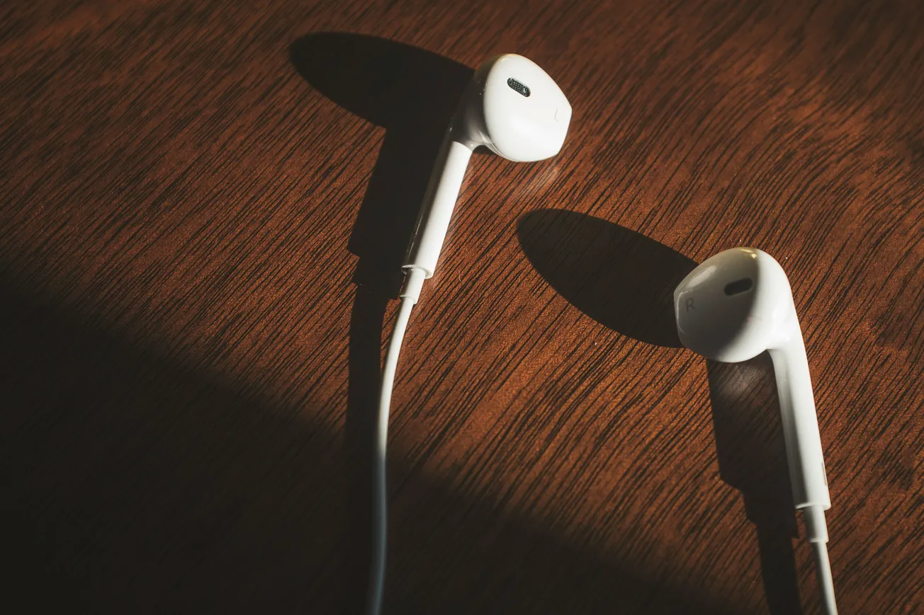 White earbuds lie on a wooden surface, lit by warm sunlight, casting distinct shadows. The scene conveys a calm, minimalist vibe.