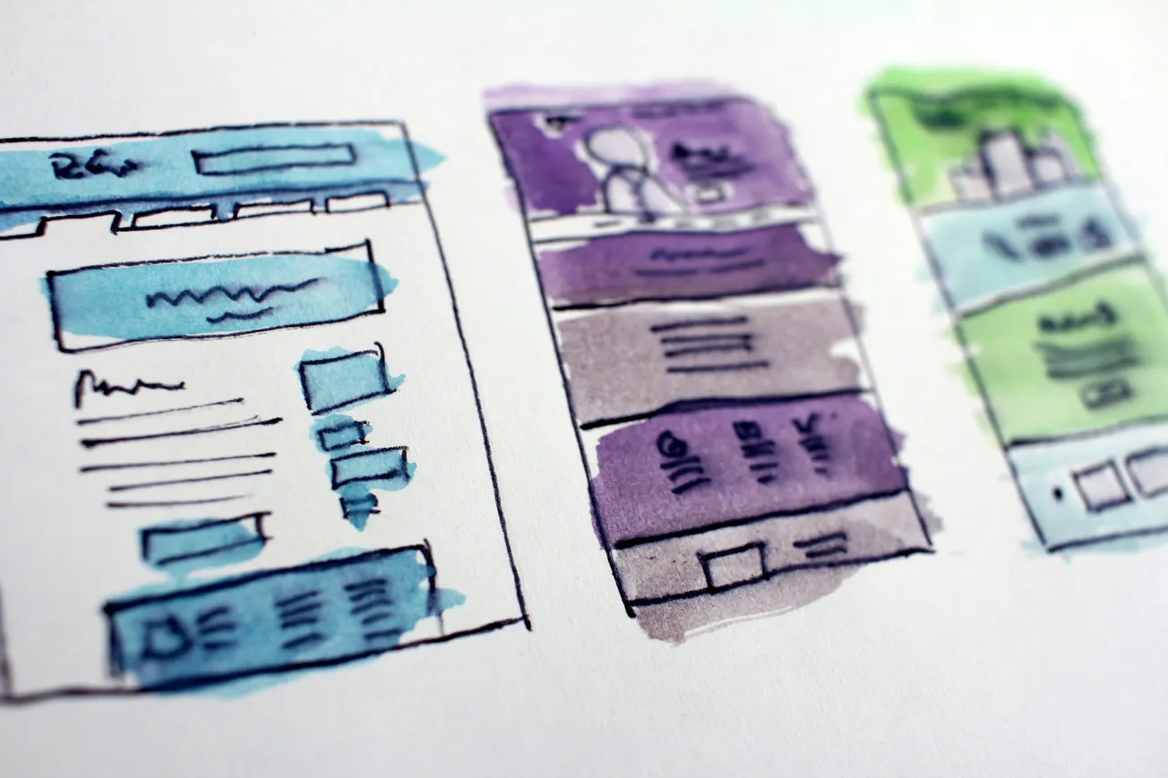 Watercolor sketches of three web page designs in blue, purple, and green on white paper. Each design shows text boxes and icons, conveying creativity.