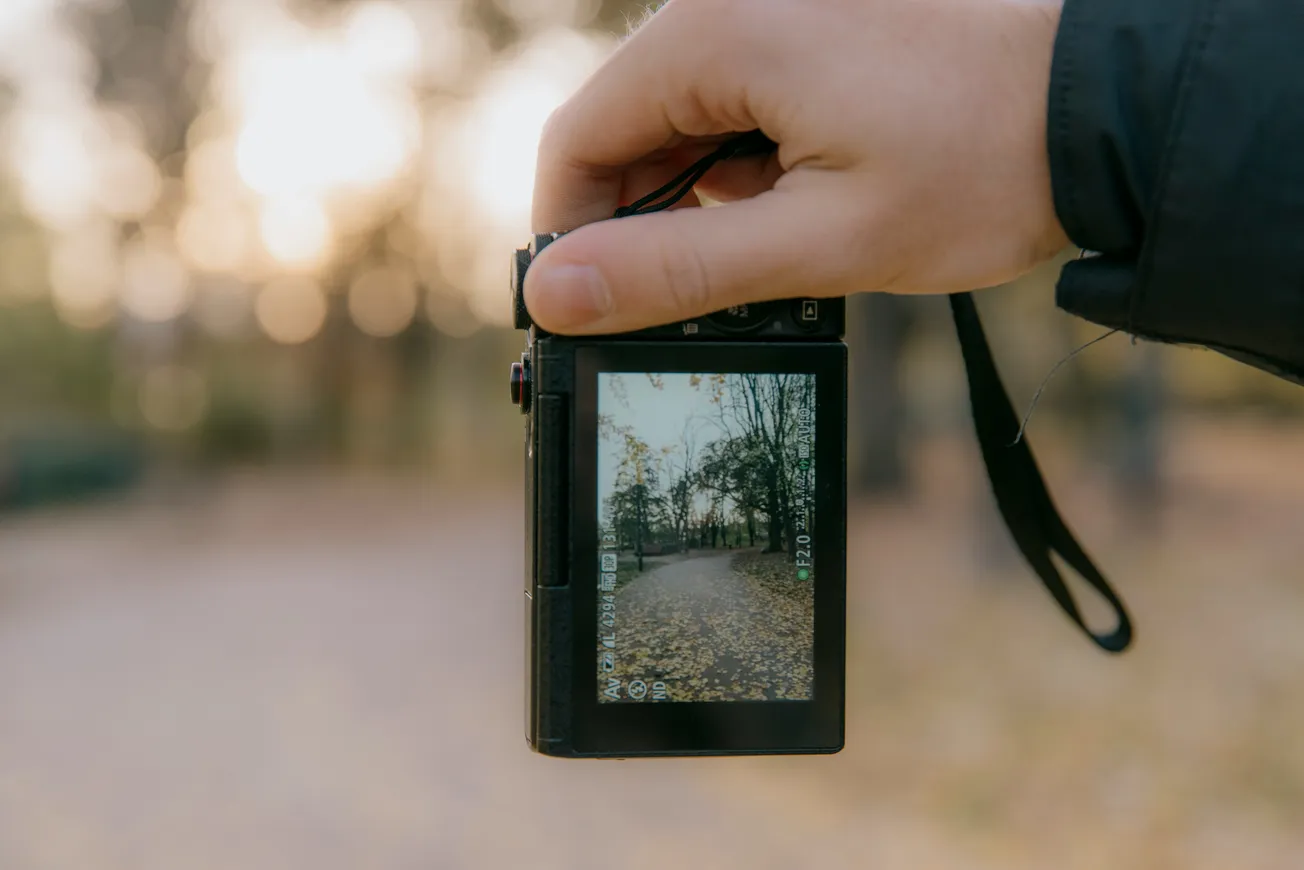 A hand holds a digital camera displaying a wooded path covered in autumn leaves. The background shows a soft-focus, sunlit forest, evoking a serene atmosphere.