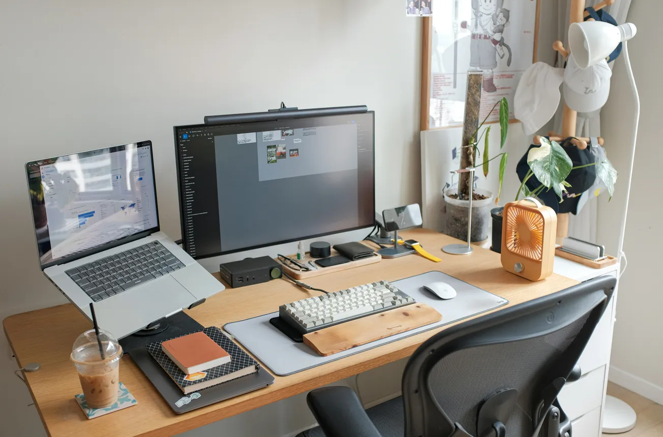 Neat workspace with a laptop, large monitor, keyboard, notebooks, and a mouse on a wooden desk. Nearby are a plant, lamp, iced coffee, and small fan.