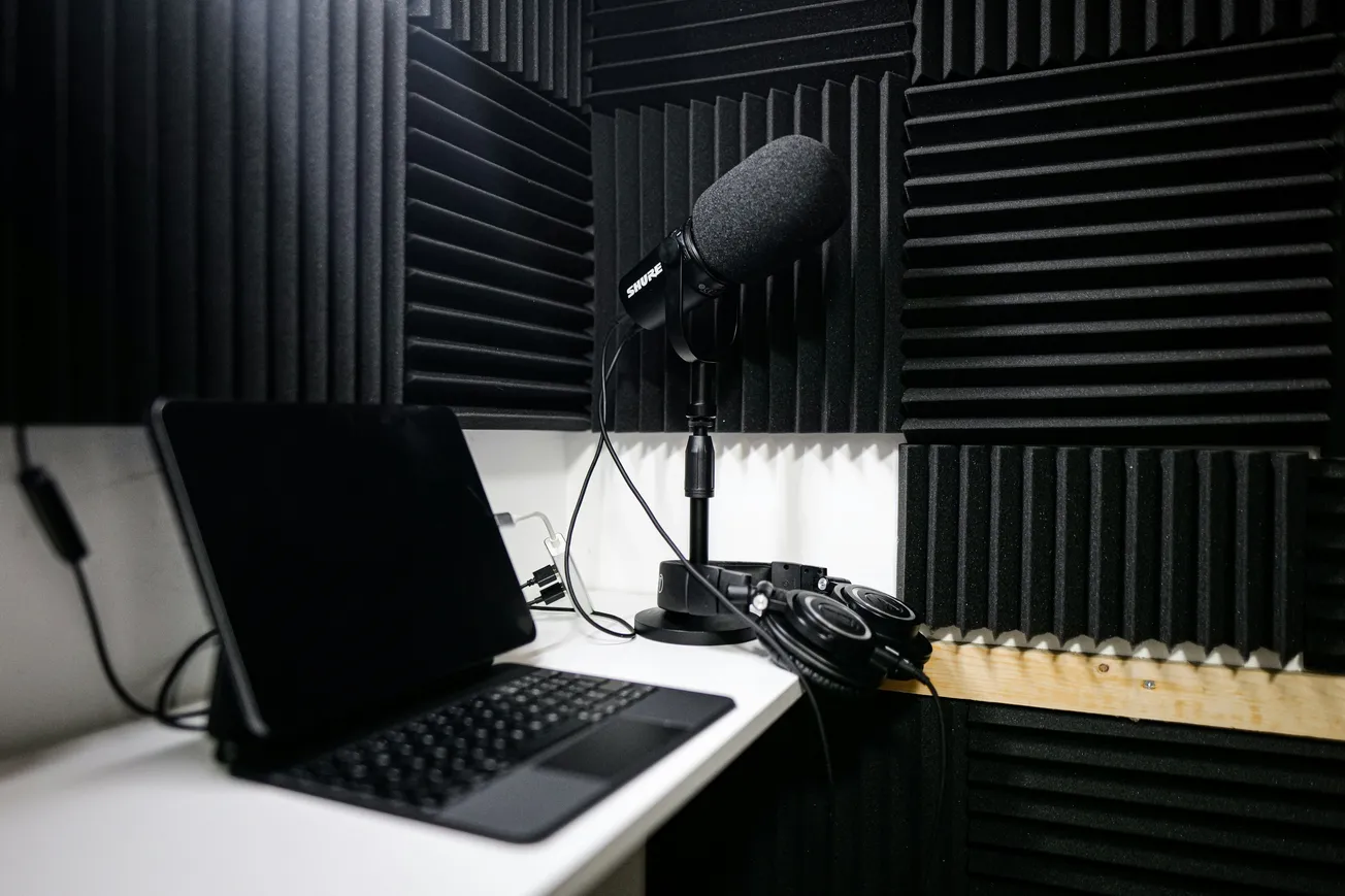 A recording studio with soundproofing foam on walls, featuring a microphone on a stand, a laptop, and headphones on a desk, creating a focused atmosphere.