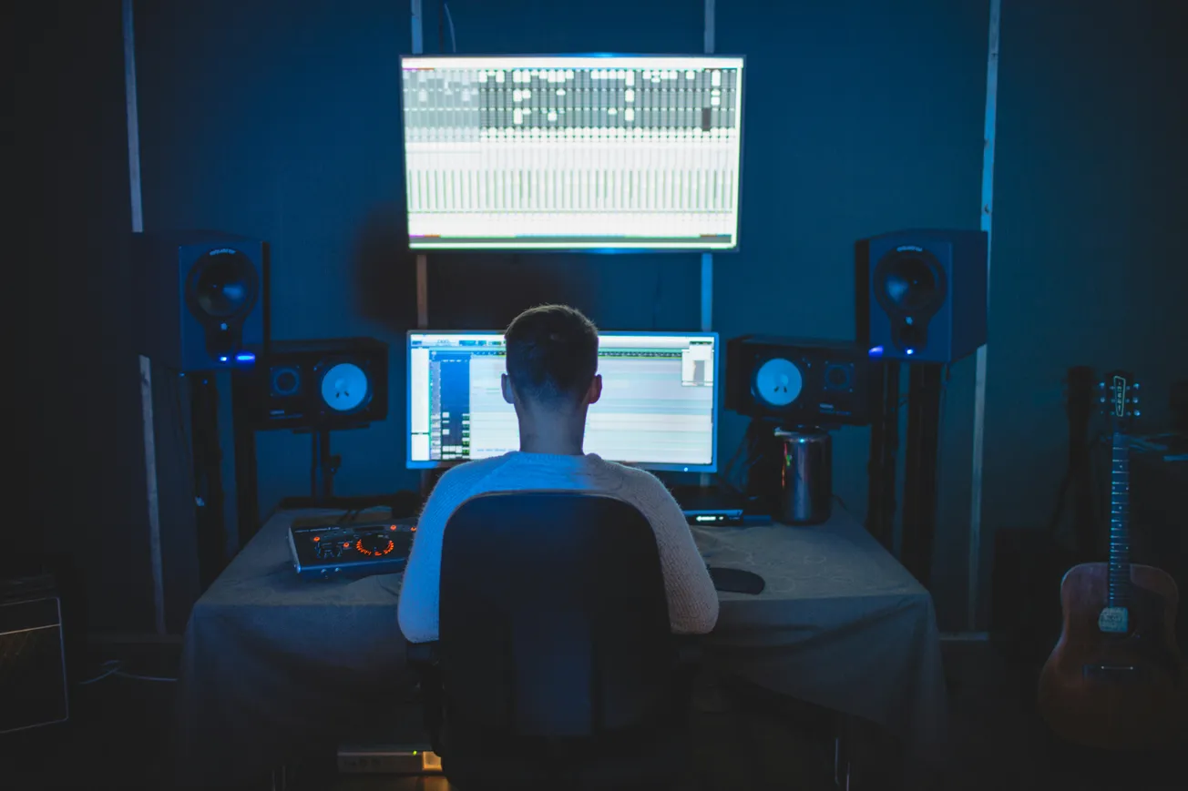 A person sitting in a dimly lit recording studio with dual monitors displaying audio editing software. Surround sound speakers and a guitar are visible.