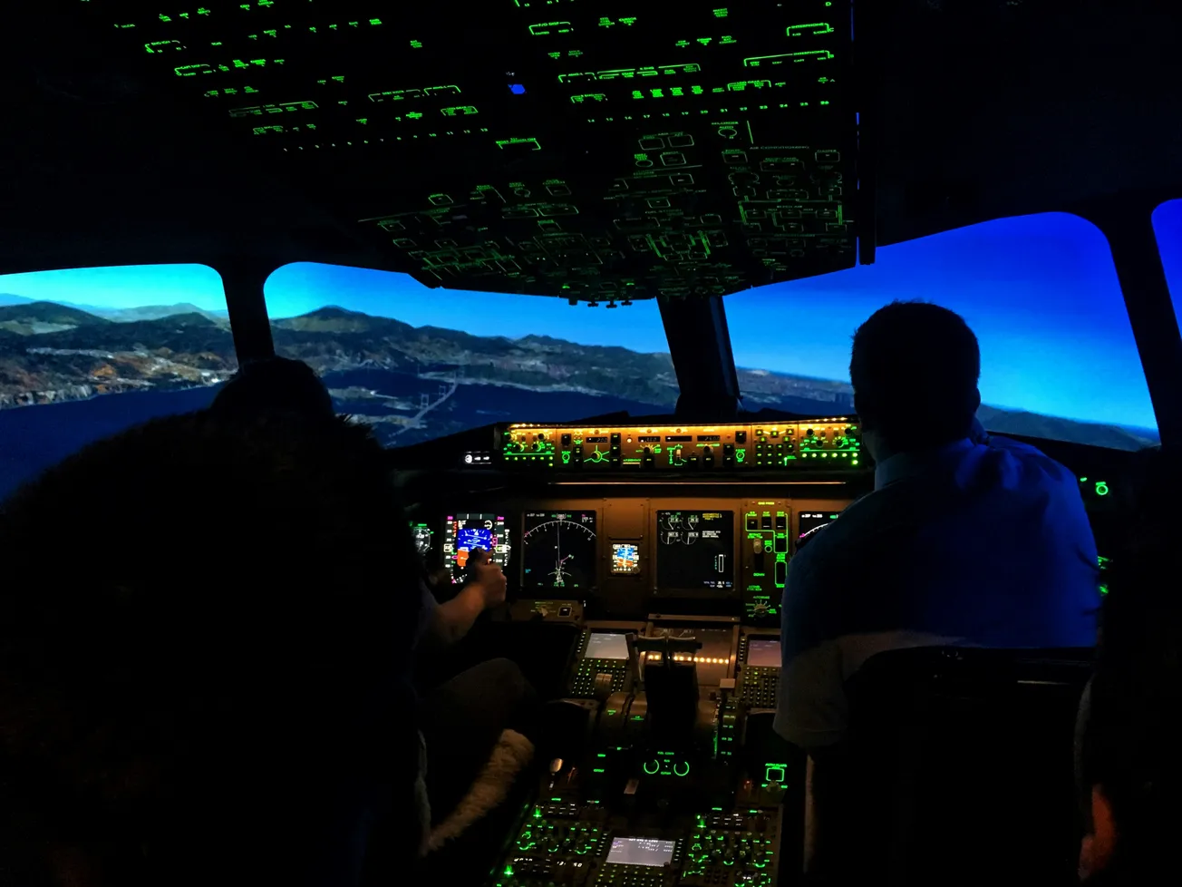 Flight simulator cockpit with pilots at controls. Electronic panels glow green, simulating a flight over mountainous landscape under a dark blue sky.