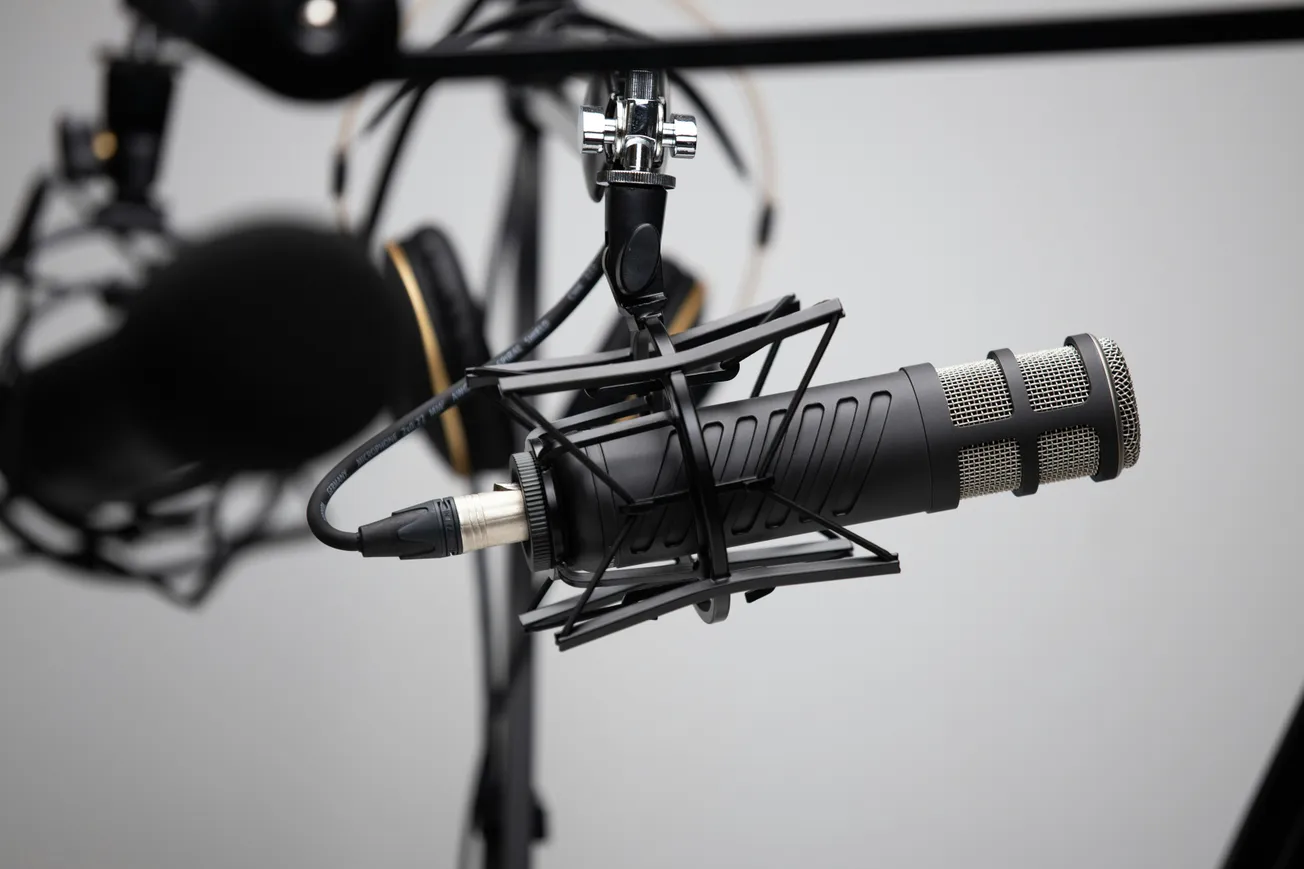 Close-up of a professional microphone in a shock mount, with blurred headphones in the background. The setting feels technical and focused.