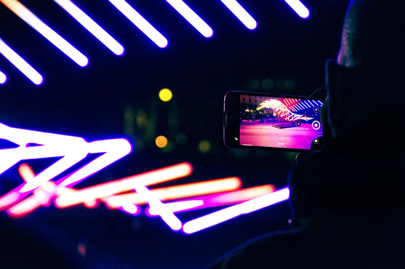 A person captures bright purple and pink neon lights with a smartphone. The scene is vibrant, glowing against a dark background, creating a dynamic, modern feel.