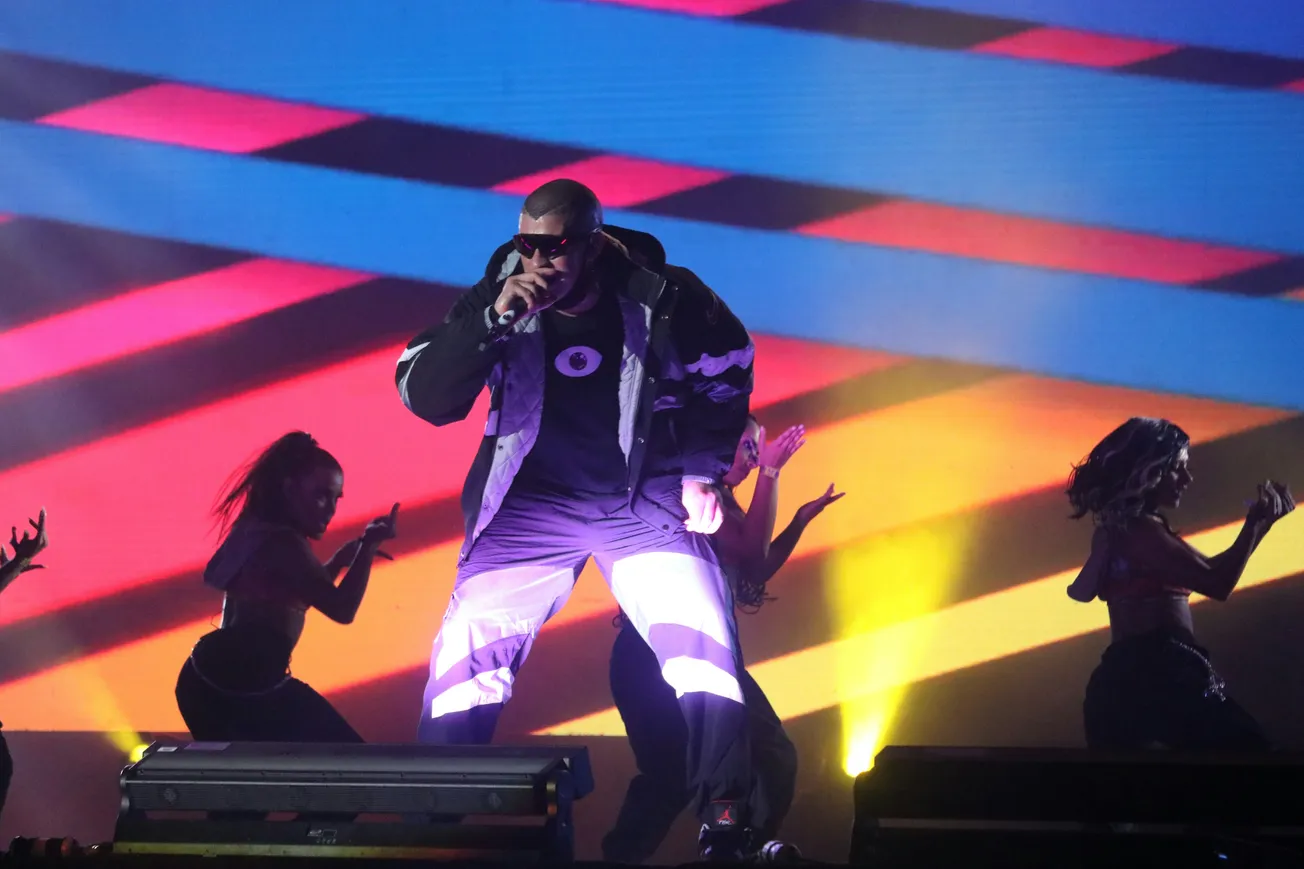 Bad Bunny in a jacket and sunglasses sings into a microphone on stage, surrounded by dancers. Colorful diagonal lights create a vibrant backdrop.
