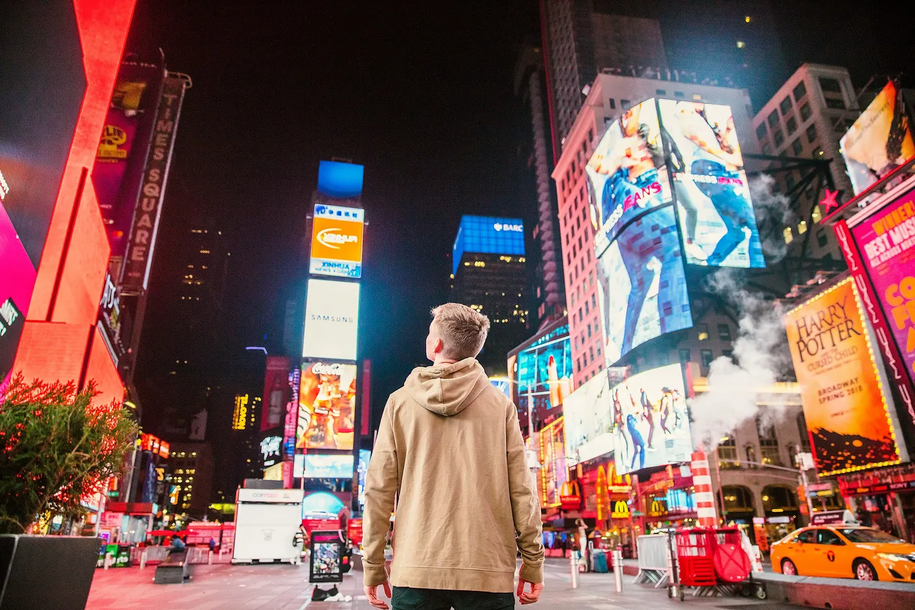 A person in a beige hoodie stands in brightly lit Times Square at night, surrounded by colorful billboards and bustling city lights, conveying a sense of wonder.
