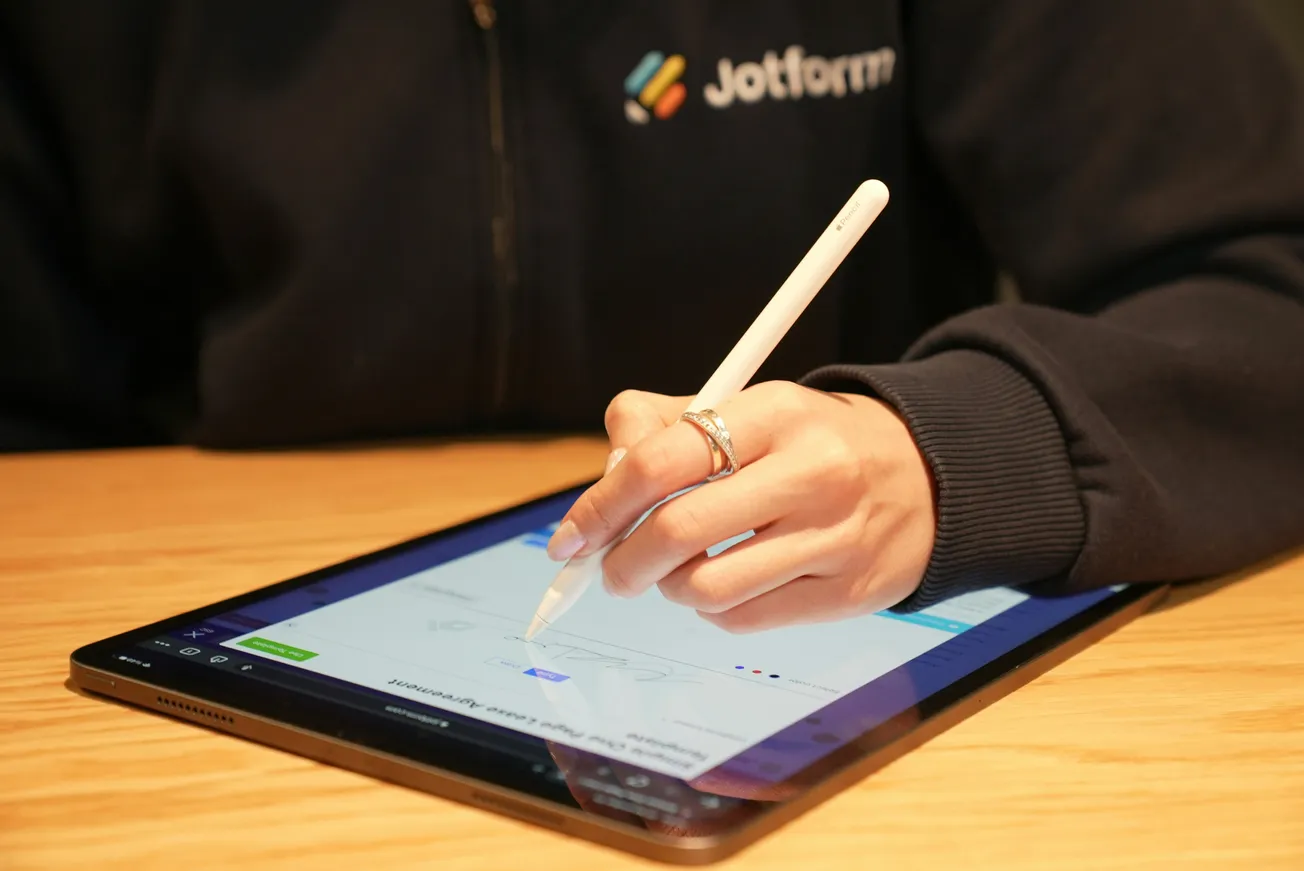 A person wearing a dark jacket uses a stylus to write on a tablet screen atop a wooden surface. The jacket features a Jotform logo.