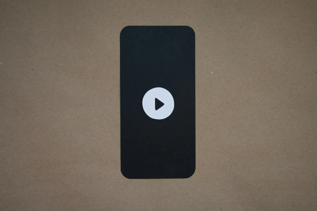 A black rectangle with rounded corners on a brown background features a white play button in the center, resembling a smartphone screen.