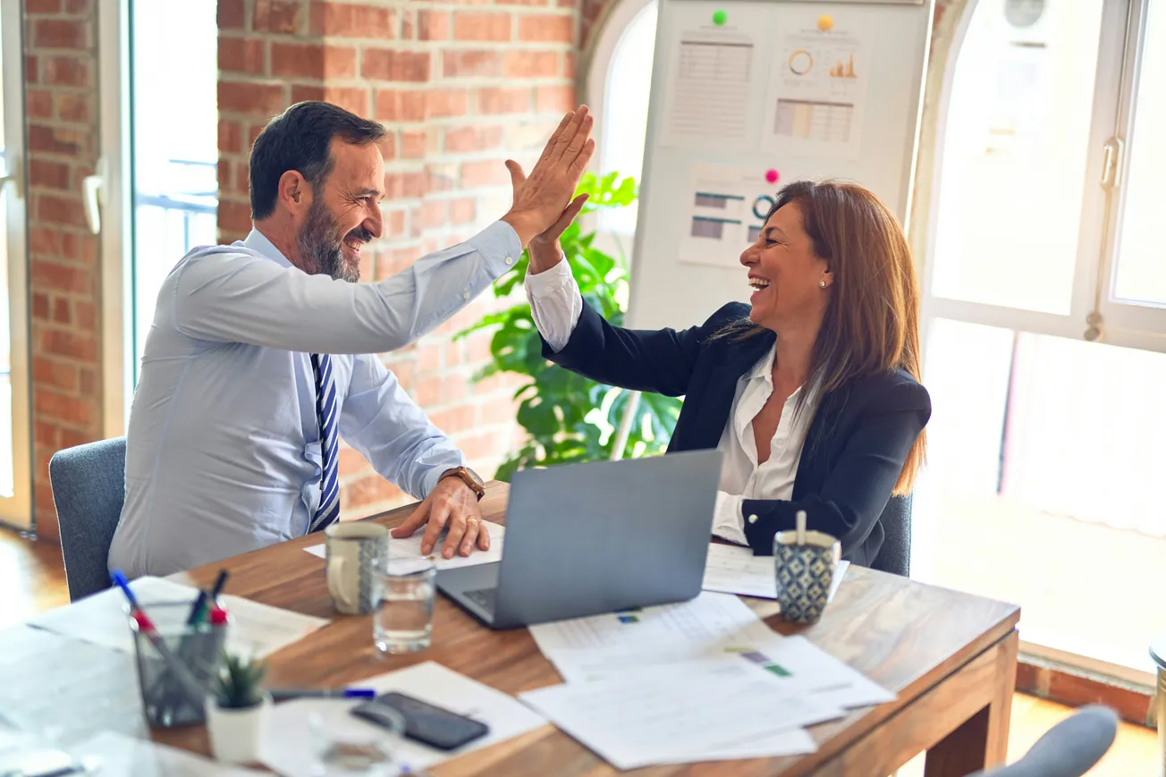 A man and woman, both in business attire, share a joyful high-five at a wooden desk with a laptop and papers, conveying teamwork and success.