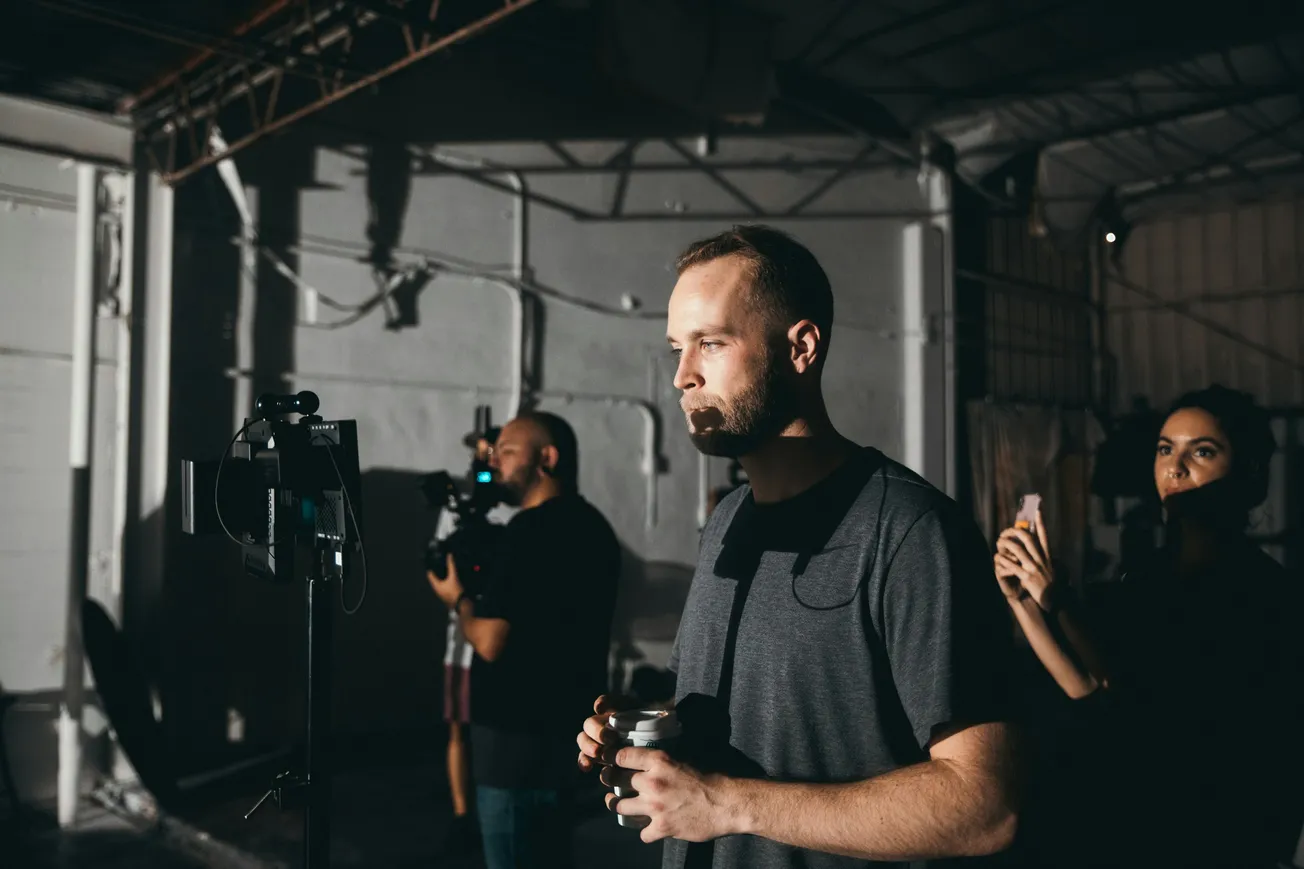 Man in a dimly lit room holds a coffee cup, focused on a monitor. Two others, one with a camera, work in the background. Industrial setting, serious tone.