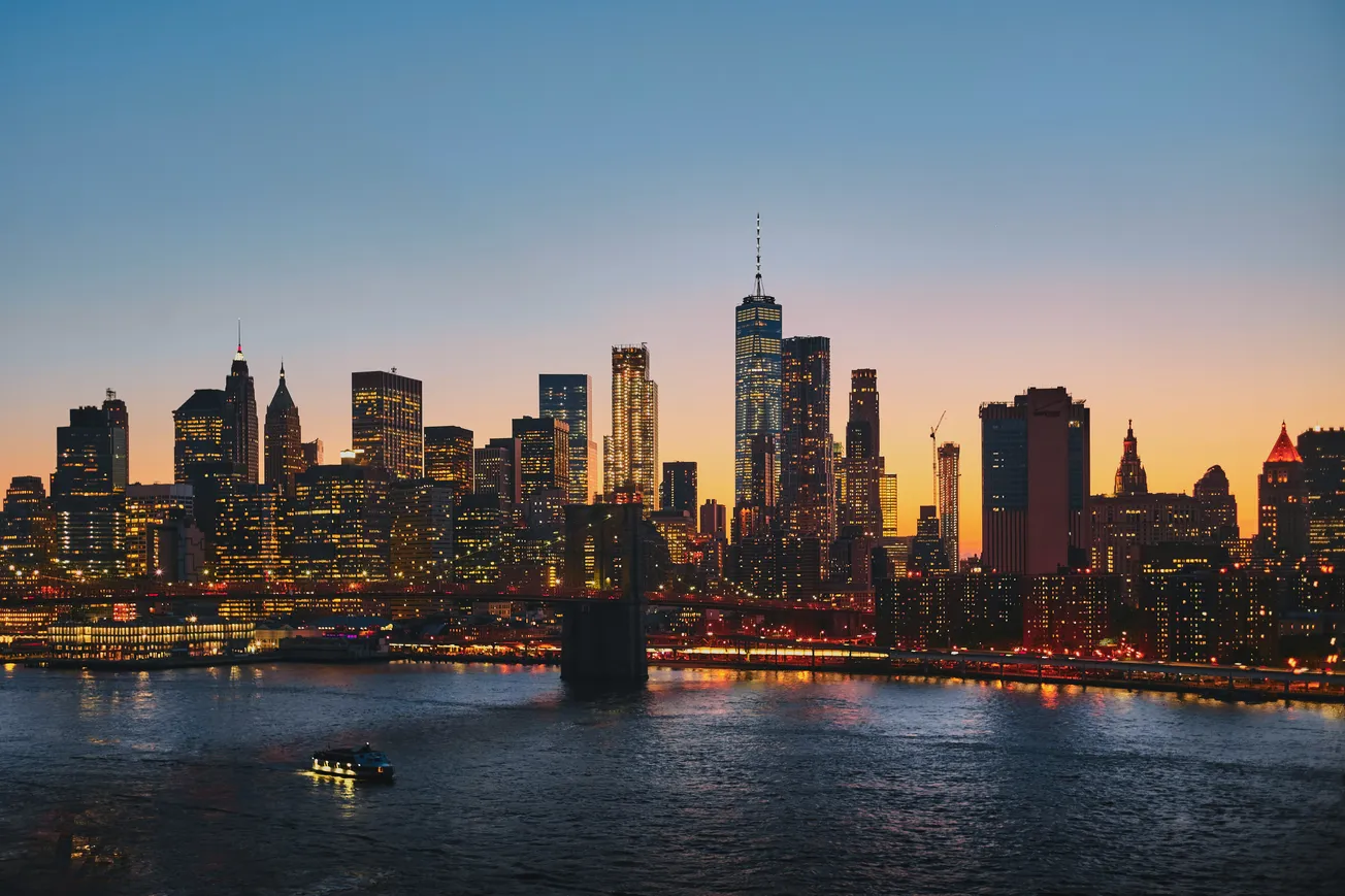 New York City skyline at dusk, with illuminated skyscrapers under a gradient twilight sky, reflecting on the calm river below, conveying a serene urban vibe.