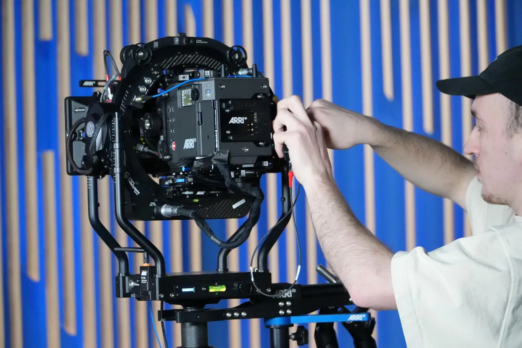 A person wearing a cap adjusts a large ARRI movie camera mounted on a complex rig. The background features vertical blue and beige stripes, creating a modern and technical atmosphere.
