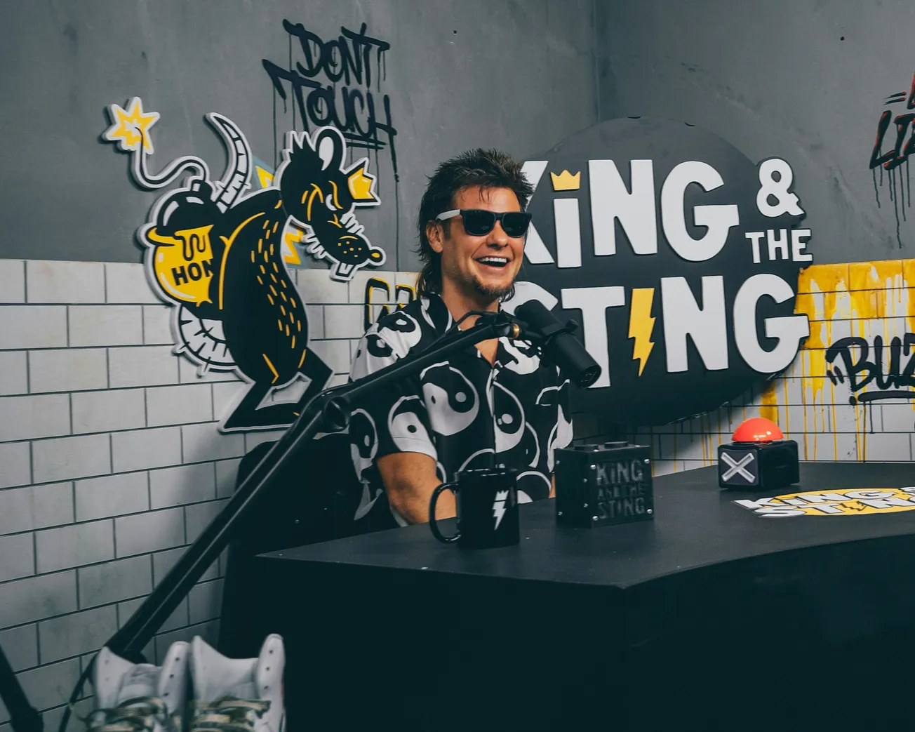 Theo Von in sunglasses sits at a podcast desk with a black and white yin-yang shirt. The background has graffiti-style art and "King & the Sting" text. Playful vibe.