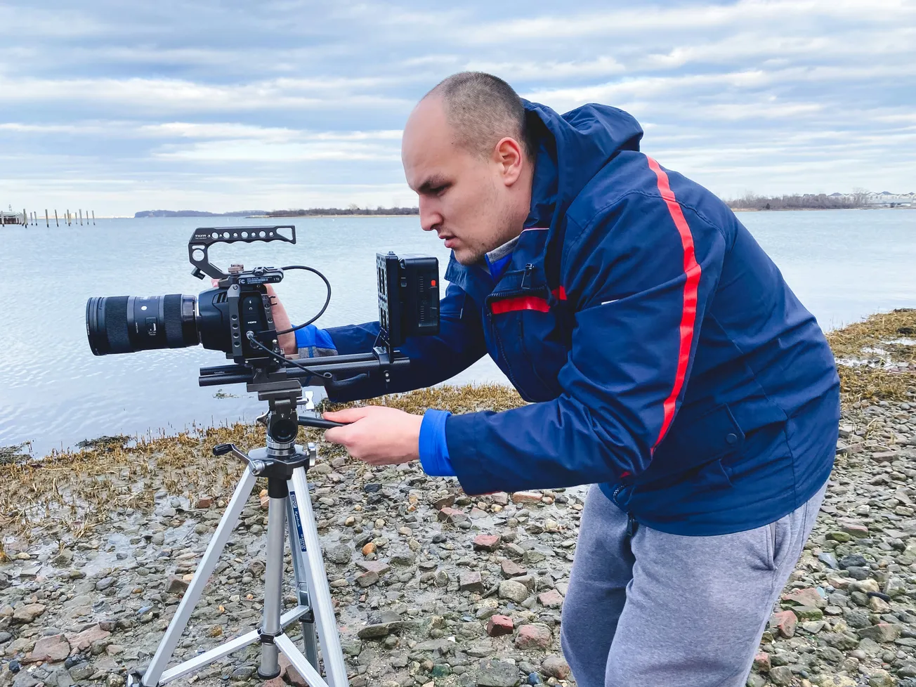 A man in a blue jacket uses a tripod-mounted camera on a rocky lakeside. The overcast sky sets a focused mood during the shoot.