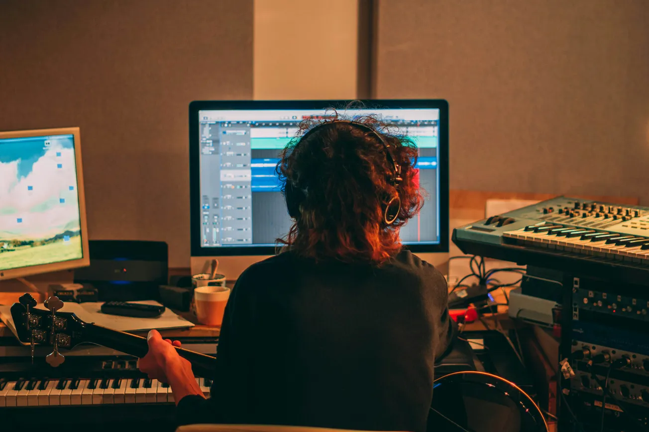 A person with curly hair wearing headphones sits at a desk with a computer monitor displaying music editing software, surrounded by a keyboard and audio equipment.