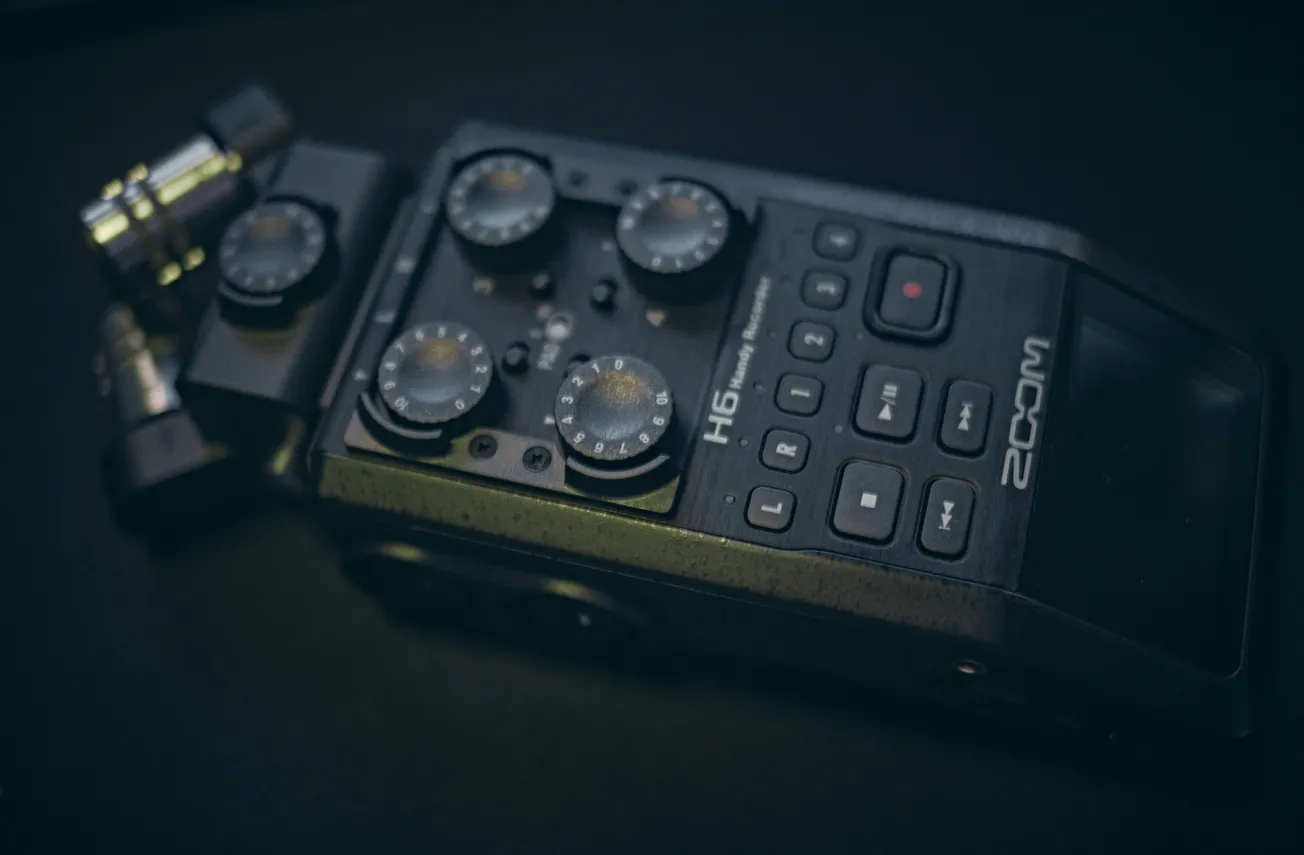 Close-up of a Zoom H6 Handy Recorder on a dark surface. The device features four round control knobs and multiple buttons, conveying a sense of professional audio equipment.