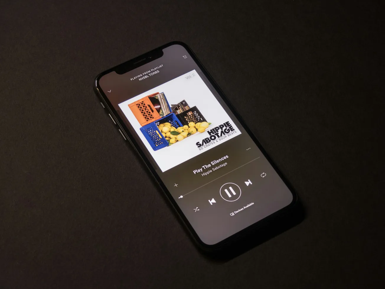Smartphone on a dark surface displaying a music streaming app. The album cover shows crates filled with lemons. Minimalist and relaxed tone.