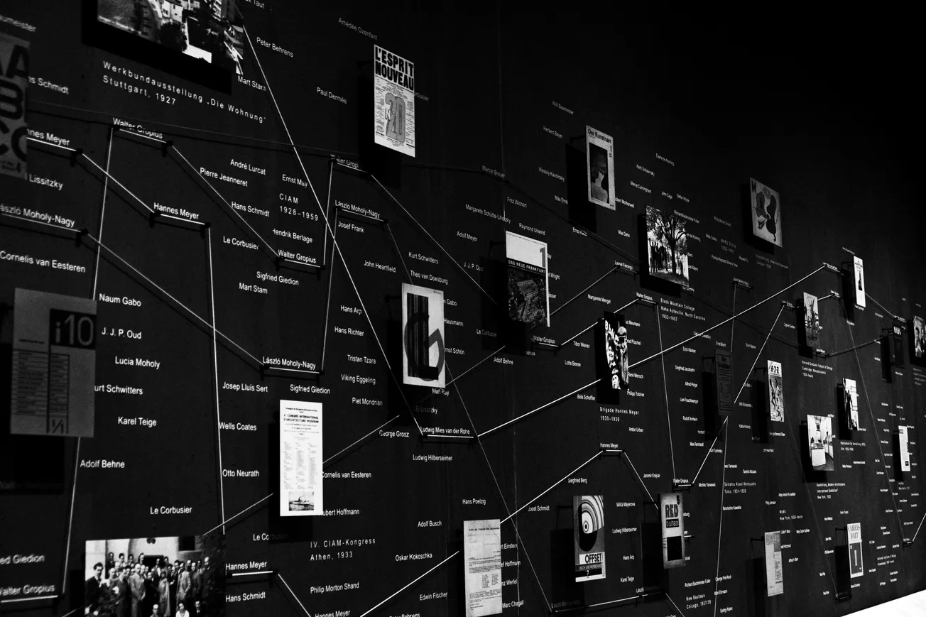 Black and white photo of a museum exhibit wall featuring interconnected artworks and texts linked by strings, suggesting a network of ideas or history.