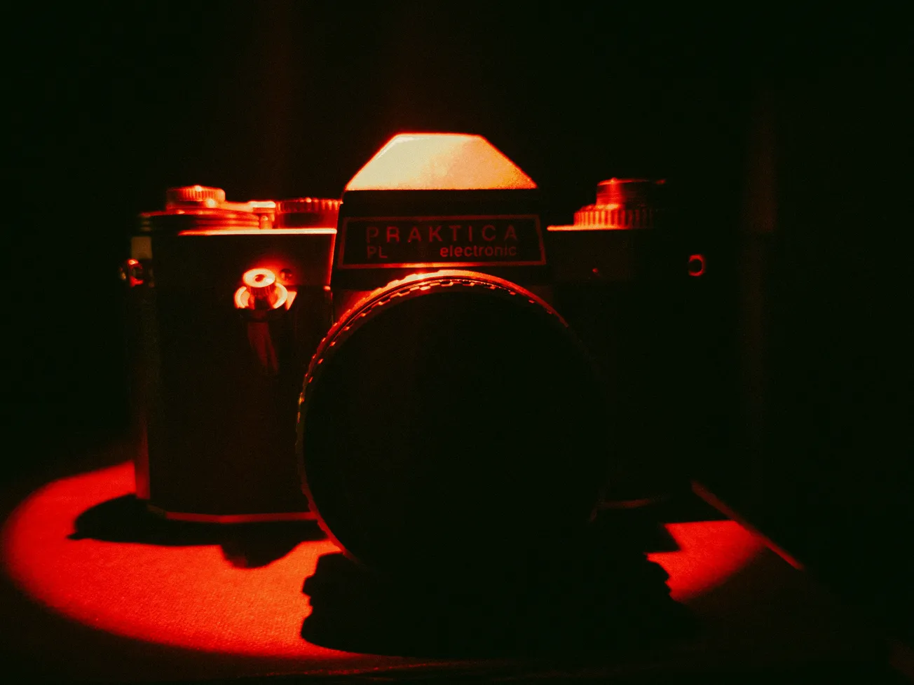 A vintage Praktica camera is bathed in dramatic red lighting, highlighting its silhouette and creating a mysterious, nostalgic atmosphere.