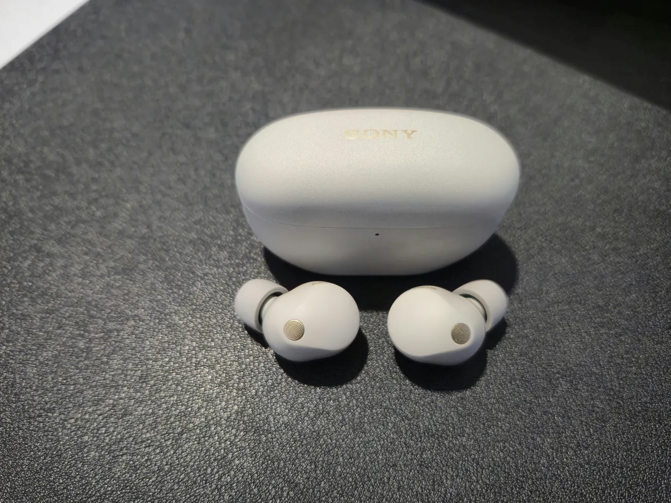 White earbuds with a matching charging case are placed on a dark textured surface. The earbuds have a small gold accent, and the lighting is soft.