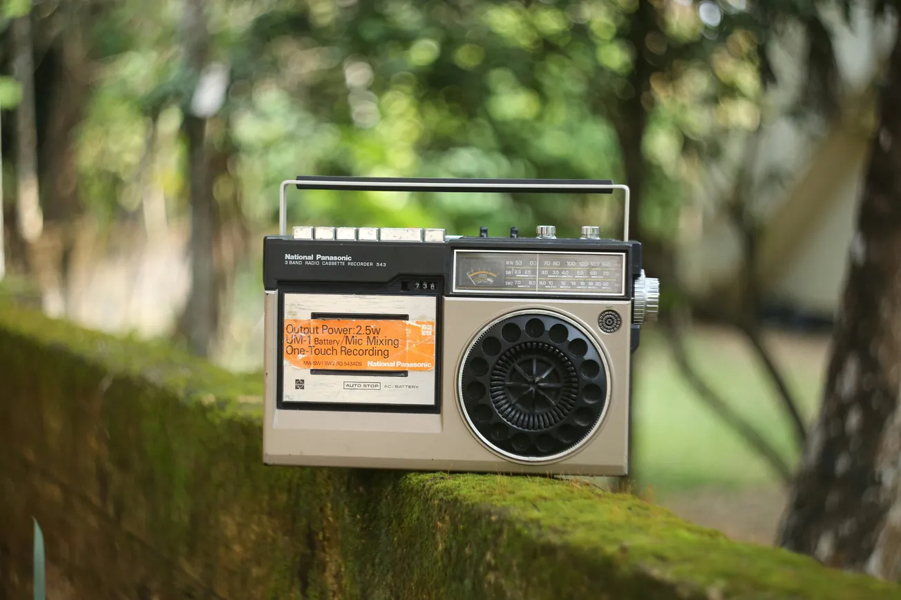 A vintage radio with dials and a large speaker sits on a mossy stone ledge. The background features blurred green trees, evoking nostalgia.