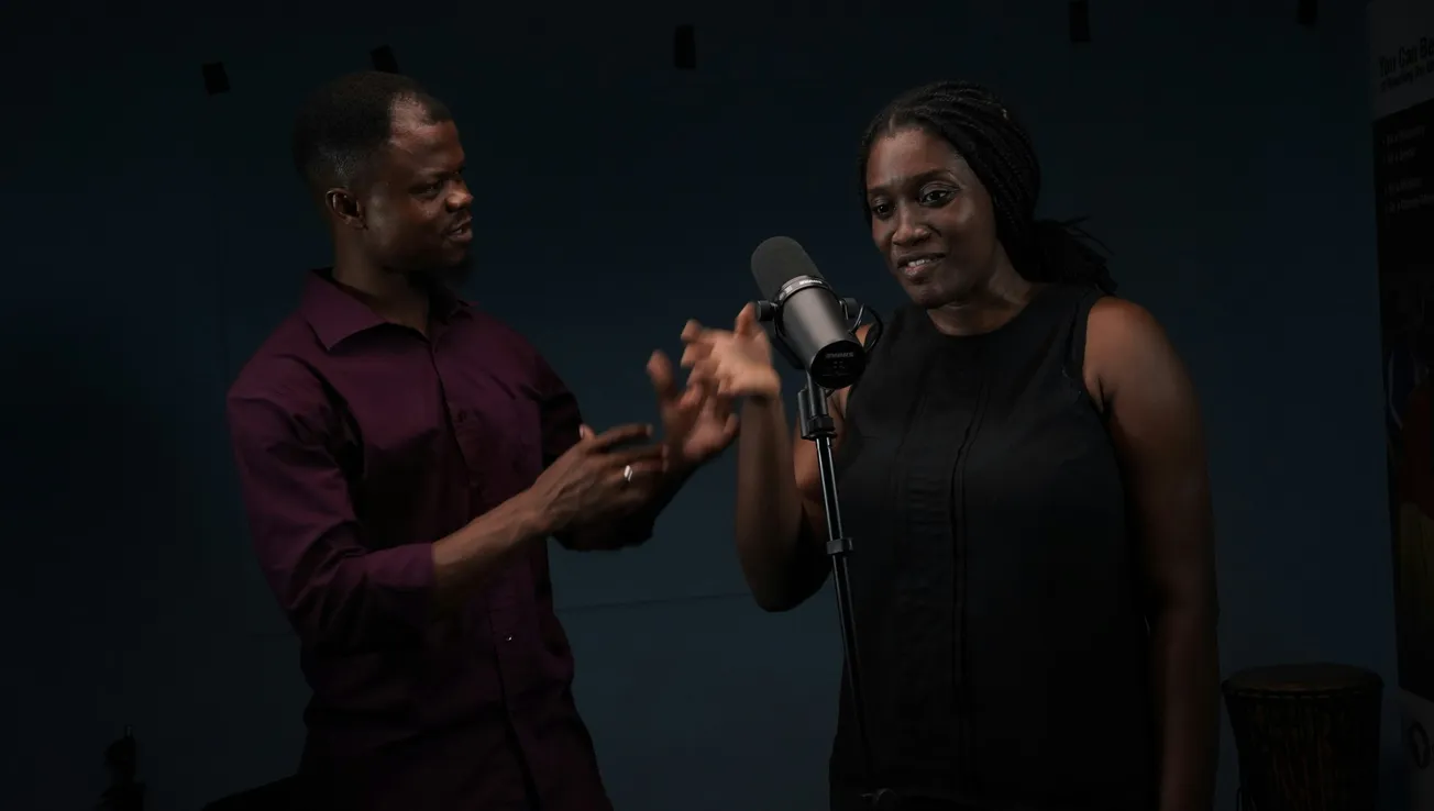 A man and woman stand in a dimly lit room with a microphone. The man gestures expressively while the woman listens, creating a focused and serious mood.
