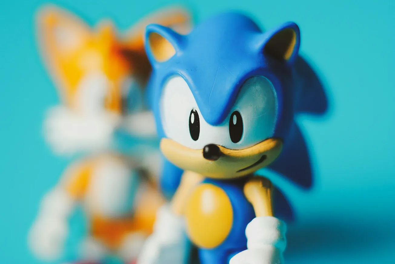 Toy figures of two animated characters are shown. In the foreground, a blue character with large eyes smiles slightly. Behind, a blurred orange character. The background is blue.