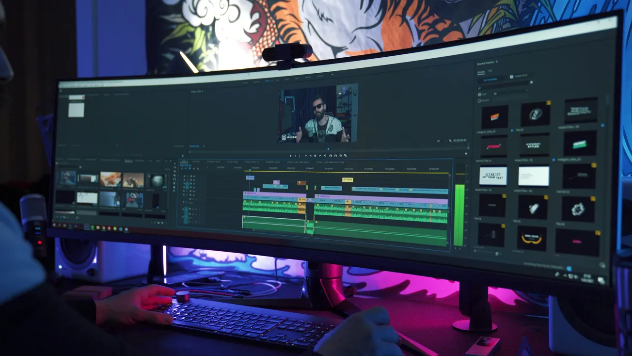 A person works at an ultra-wide monitor showing a video editing software interface with multiple colorful timelines. The room is dimly lit with a vibrant mural.
