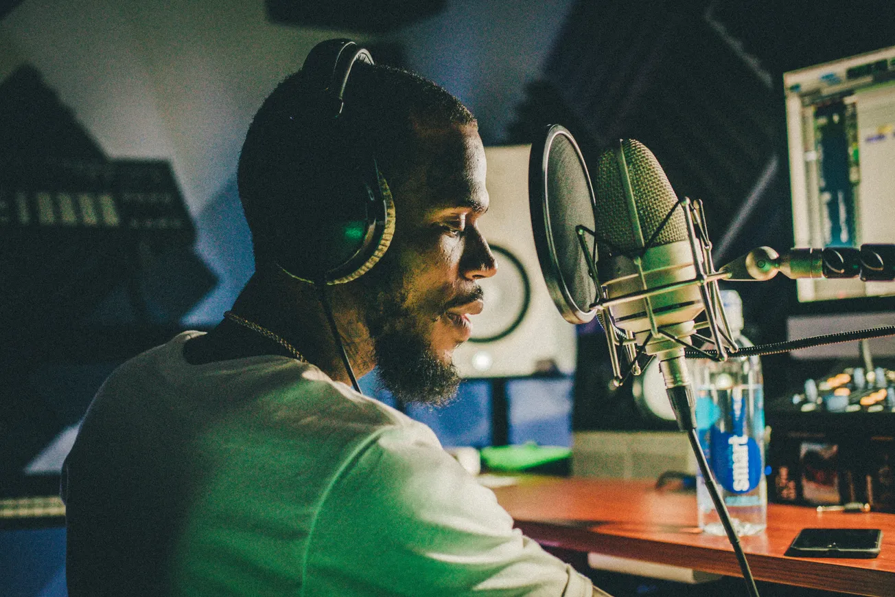 A man in headphones at a recording studio is focused on a microphone, with monitors and soundproofing in the background, suggesting a creative atmosphere.